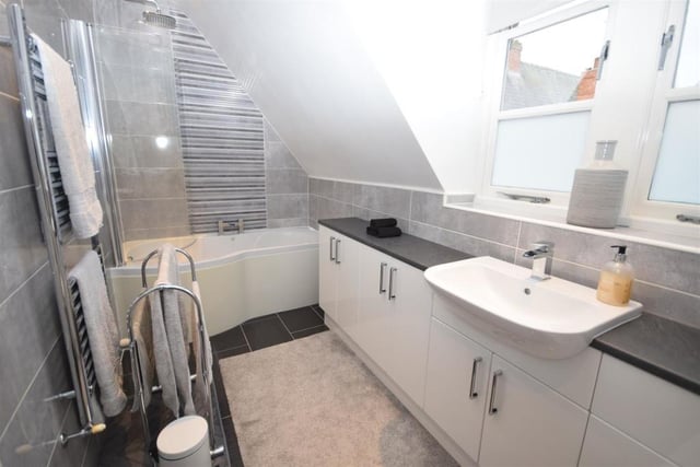 This contemporary en suite serves the main bedroom. It is fitted with a P-shaped, panelled bath that has a rainfall shower above, a low-flush WC and a wash basin in a vanity unit. The floor and walls are tiled, while there is also a chrome, heated towel-rail and warm, atmospheric downlighting.